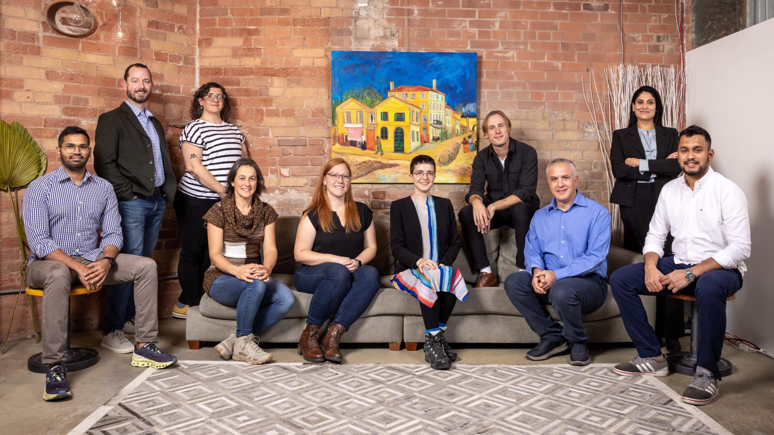 Ten people sit on and around a sofa in front of a painting on a brick wall.